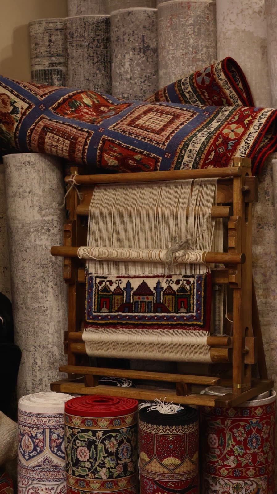A vertical loom part-way through a rug: warp threads strung under tension, a few centimetres of knotted pattern completed at the base, and rolled rugs stacked behind.